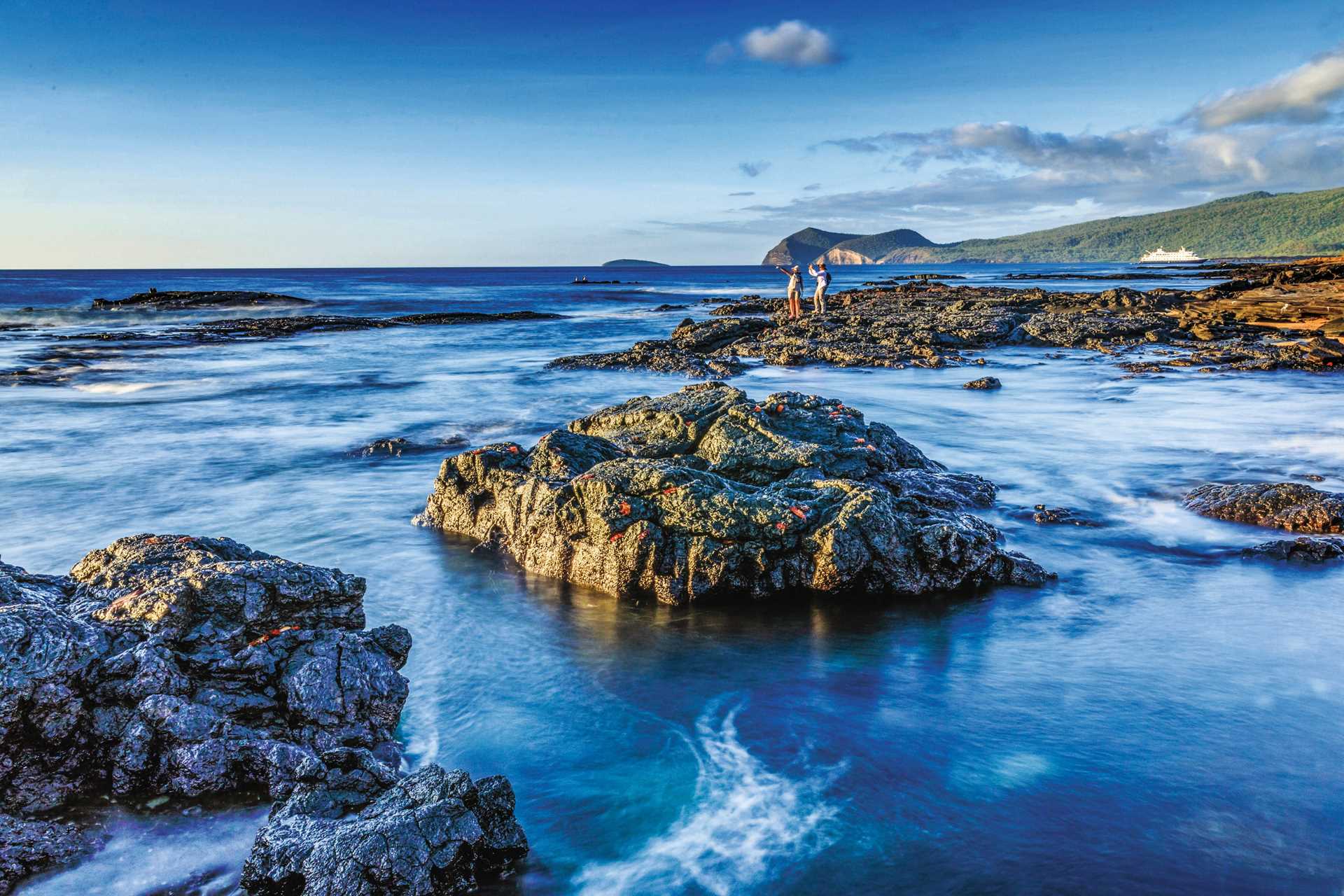 Two guests in the distance takes photos while walking on lava-formed rocks on Santiago Island, Galápagos.